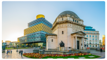 The Library of Birmingham with its modern patterned design and golden cylindrical top standing behind the historic Hall of Memory war memorial, surrounded by flowerbeds and a paved square with people walking.