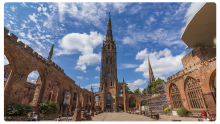 Ruins of Coventry Cathedral in England, showing the tall spire and red sandstone walls under a bright blue sky with scattered clouds, with visitors walking through the open courtyard.