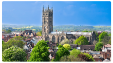 St. Mary’s Church in Warwick, England, with its tall square tower rising above the town’s red-brick rooftops, set against a clear blue sky and rolling countryside in the background.
