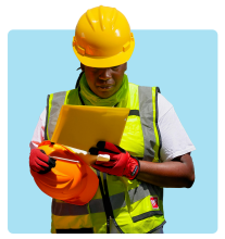 Construction worker wearing a yellow hard hat, safety vest, and gloves, holding a tablet and an orange safety helmet while checking information