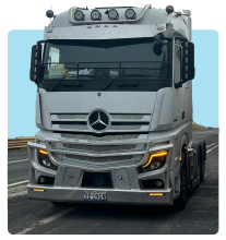 Silver Mercedes-Benz Actros truck driving on a road, showing the front view with headlights on and a prominent Mercedes logo on the grille.